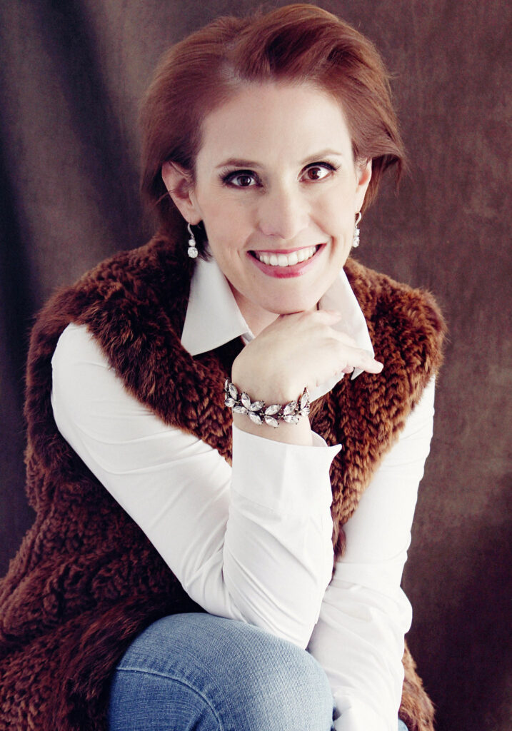 Picture of author Bettina Restrepo who has short red hair, cream colored skin and is wearing a white shirt, brown vest and blue jeans.
